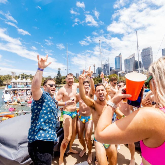 A group of people in swimwear are partying on a luxury yacht under a clear blue sky, holding drinks and cheering. City skyscrapers and yachts are visible in the background, making it a perfect example of Sydney boat party hire.