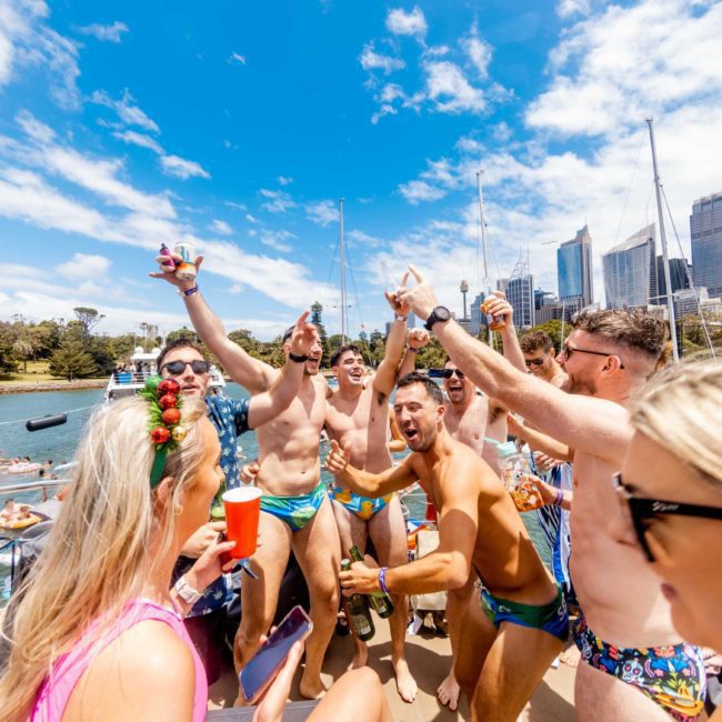 A group of people in swimwear celebrate on a boat under sunny skies with city buildings in the background. Some hold drinks, and there is a sense of cheer and excitement, perfect for corporate boat events Sydney or a vibrant Catamaran party Sydney.