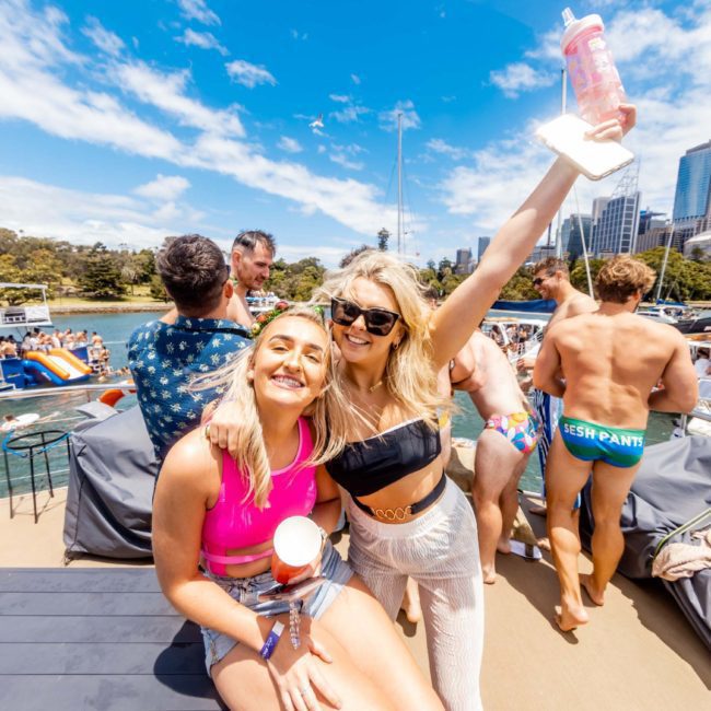 Two women smiling and posing for the camera on a boat, with one raising her arm holding a drink. Several other people and boats are visible in the background, enjoying a sunny day. Perfect setting for a luxurious catamaran party in Sydney Harbour.