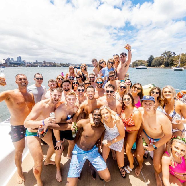 A large group of people in swimsuits stands on a luxurious yacht, posing and smiling for a photo with a cityscape and the Sydney Harbour Bridge in the background.