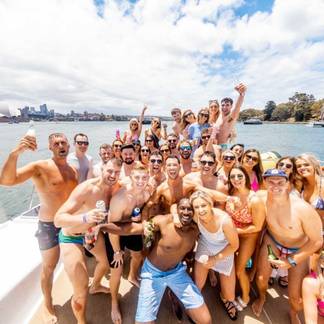 A group of people in swimwear are posing and smiling on a boat with drinks, enjoying a Sydney boat party hire. They are on a sunny waterway with the city's landmarks in the background.
