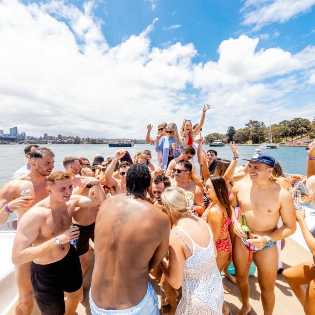 A large group of people in swimwear are celebrating on a boat under sunny skies, with a cityscape and landmarks visible in the background. Consider Sydney boat party hire to make your event stand out!