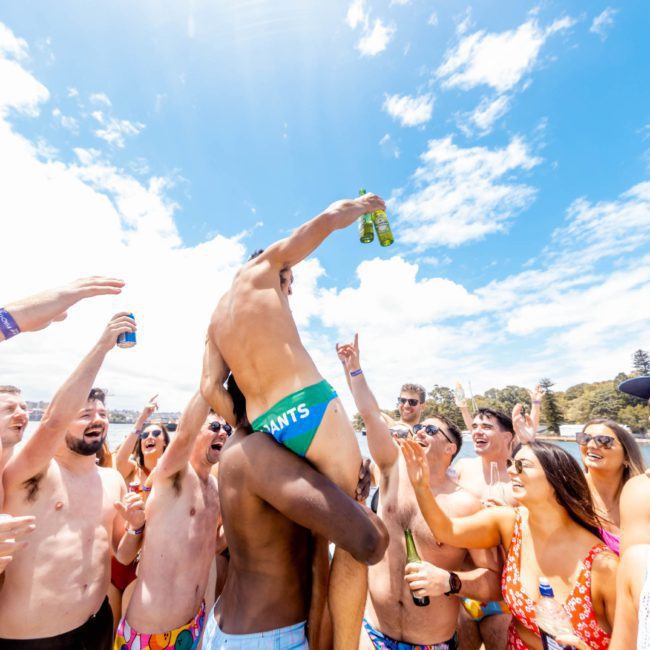 A group of people in swimwear celebrating outdoors under a sunny sky on a catamaran party in Sydney. One person is lifted above the crowd holding a drink, while others cheer and raise their own beverages.