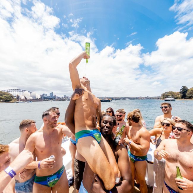 A group of men in swimwear celebrates on a private yacht charter Sydney Harbour, with one person being lifted and drinking from a bottle. The scene is set on a sunny day with a cityscape and a bridge in the background.