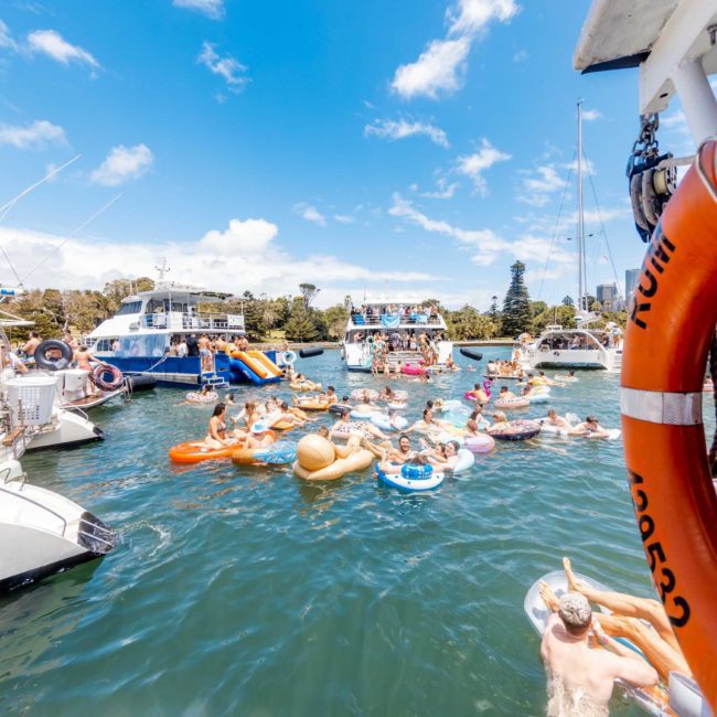 People enjoying a sunny day on the water, floating on various inflatables and surrounded by boats. A lifebuoy is visible in the foreground, while others look toward a luxury yacht hire Sydney service nearby.