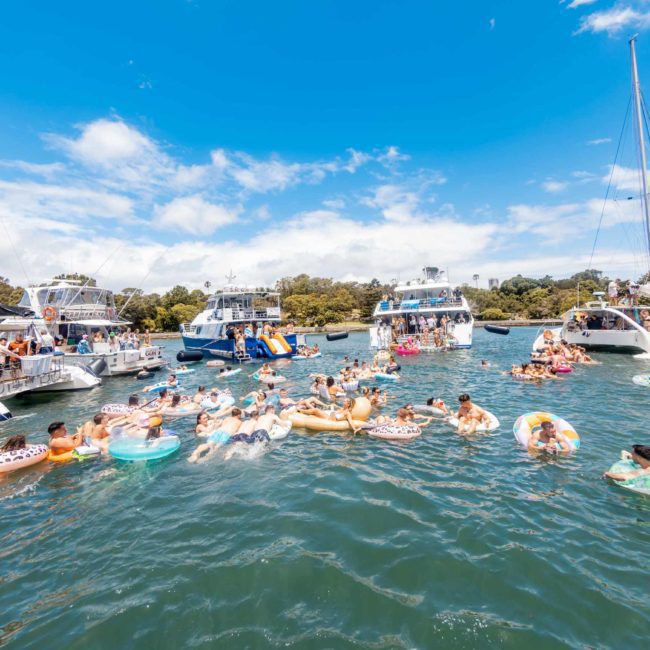 People enjoying a sunny day on the water, swimming and floating on inflatable rings, surrounded by anchored boats with slides and other recreational activities. Consider DJ boat hire Sydney for added fun or luxury yacht hire Sydney to elevate your experience on the water.