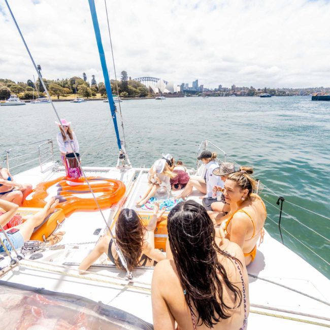 A group of people in swimwear enjoying a sunny day on a sailboat. Some are standing, while others sit or lie on pool floats. The background includes a calm body of water, other boats, and a distant cityscape, perfect for Sydney boat party hire and corporate boat events in Sydney.
