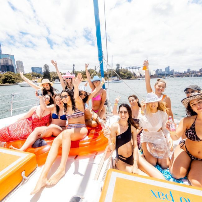 A group of people in swimwear are sitting and standing on a luxury yacht hire Sydney in a sunny harbor with a city skyline in the background, smiling and raising their arms in celebration.