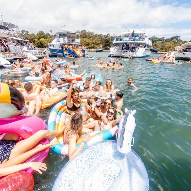 A large group of people enjoy a sunny day on the water, lounging on various inflatable floats, with boats moored nearby. The scene is lively and filled with colorful inflatables, perfect for a catamaran party in Sydney.