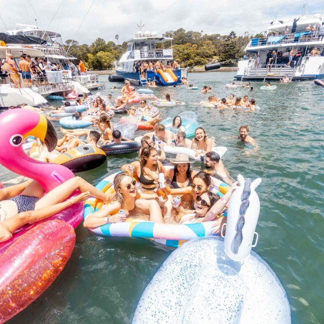 A group of people float on inflatable toys in the water surrounded by boats, enjoying sunny weather and the vibrant atmosphere of a DJ boat hire in Sydney.