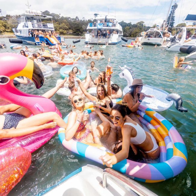A group of people enjoying a sunny day on the water with various inflatables, boats in the background, some people lounging and others in a kiddie pool, all in swimwear. Consider enhancing your experience with a luxury yacht hire Sydney for an unforgettable outing.