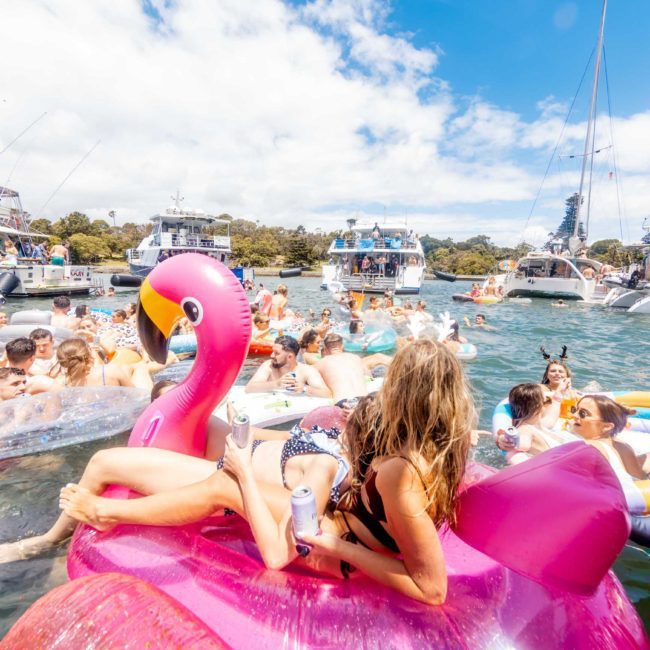 People float on inflatable pool toys, including a pink flamingo, in a crowded area of a lake or marina. Boats are anchored nearby as people enjoy a sunny day on the water, while others opt for luxury yacht hire Sydney for an upscale experience.