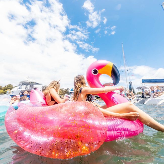 Two people are riding a large pink flamingo float in the water, surrounded by other floats and boats on a sunny day, enjoying what feels like a Catamaran party in Sydney.