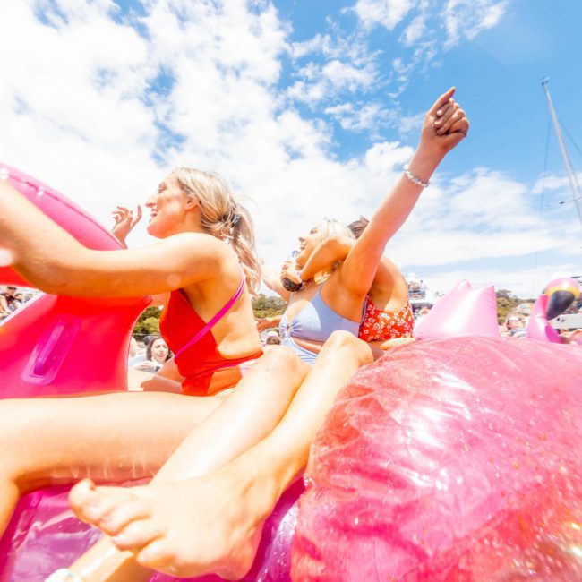 Two people in swimsuits are sitting on a large pink inflatable unicorn in a crowded water area under a sunny sky. They are surrounded by other inflatables and boats, likely enjoying the vibes of a Sydney boat party hire event.