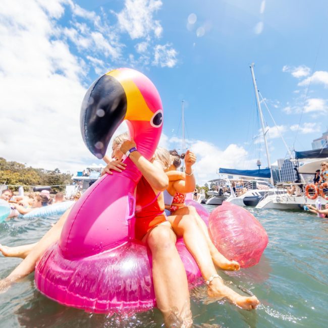 People enjoying a sunny day on the water, with one person on a large pink flamingo float, all set against the backdrop of a Luxury yacht hire Sydney.