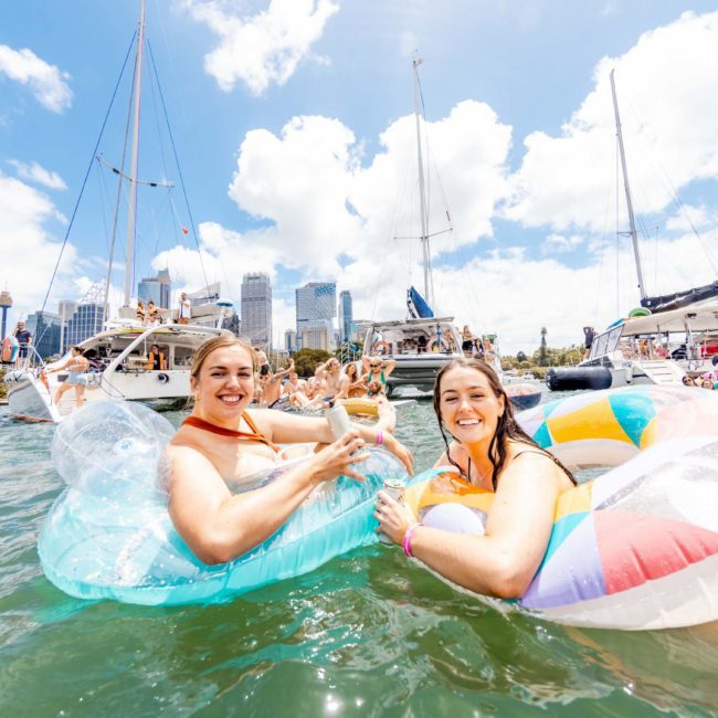 Two people float on inflatable pool chairs in a harbor, surrounded by boats and other people, with a city skyline in the background. They toast with drinks and smile at the camera, epitomizing the fun of a Sydney boat party hire.