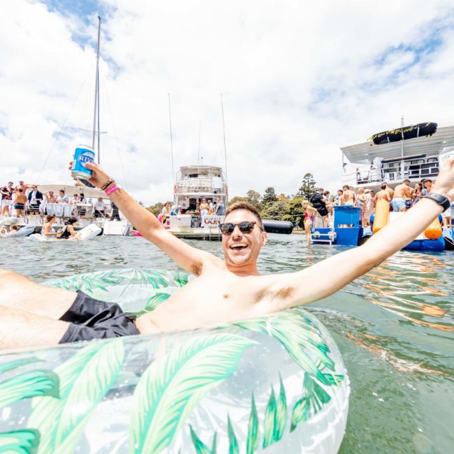 A person relaxes on a green and white float in the water, holding a can and raising their arm. A luxury yacht hire Sydney is visible among the boats and crowd in the background on a sunny day.
