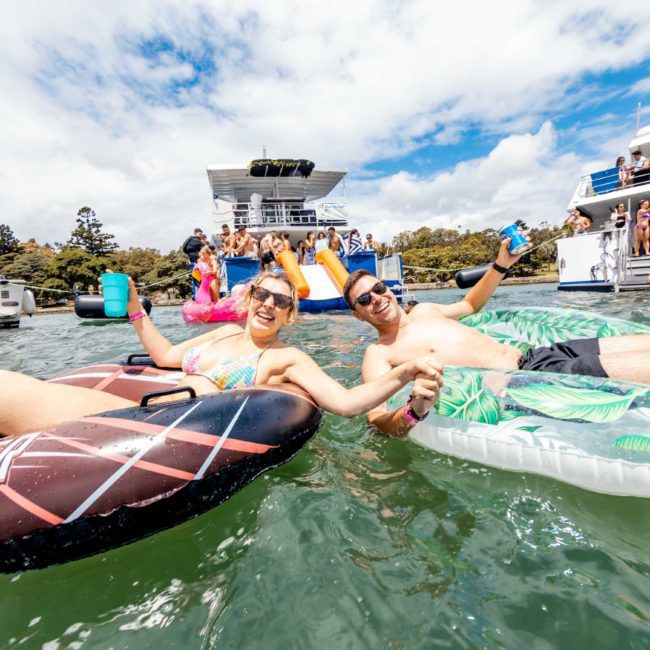 Two people hold hands while relaxing on inflatable floats in the water, surrounded by boats and people enjoying a sunny day on the lake. It's the perfect scene for a catamaran party in Sydney or even luxury yacht hire Sydney.