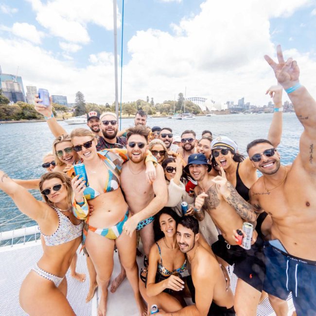 A large group of people in swimwear posing and smiling on a luxury yacht with a cityscape and water in the background on a sunny day.