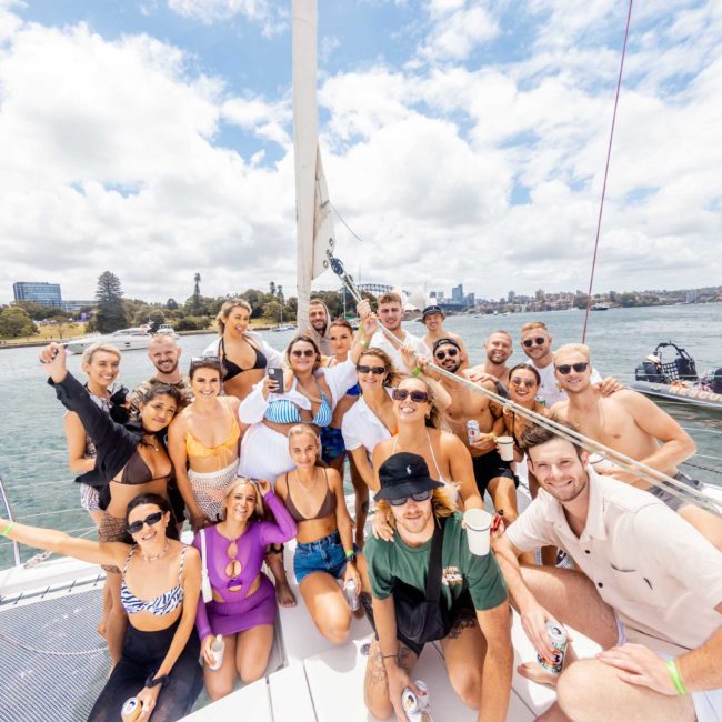 A group of people gather on a catamaran party in Sydney's harbor, smiling and posing for a photo. The background shows city buildings, trees, and other boats on the water under a partly cloudy sky.