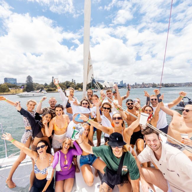 A group of people are gathered on a luxury yacht, posing for a celebratory photo. The scene is sunny with a city skyline and water in the background. They appear to be enjoying a party, typical of corporate boat events in Sydney.