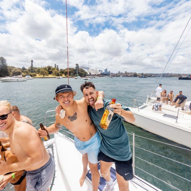 Group of friends smiling and holding drinks on a sailboat, with a scenic view of the water and boats in the background at an extravagant catamaran party in Sydney.