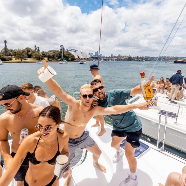A group of people enjoying a sunny day on a private yacht charter in Sydney Harbour, some holding drinks and posing for the camera. They are in swimwear with a cityscape and other boats visible in the background.
