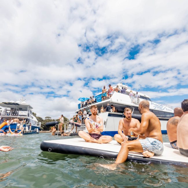 People socialize on a floating dock and boats in the water under a partly cloudy sky. Some are sitting or standing on boats, while others are in the water with inflatable rafts. It's an ideal setting for a luxury yacht hire Sydney, as everyone enjoys the beautiful day and camaraderie.