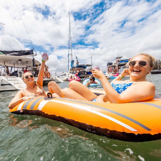 Two women on an orange inflatable raft hold drinks while floating in a marina, surrounded by boats and other people, under a partly cloudy sky. They seem to be enjoying the backdrop of a lively Sydney boat party hire with music adding to the festive atmosphere.