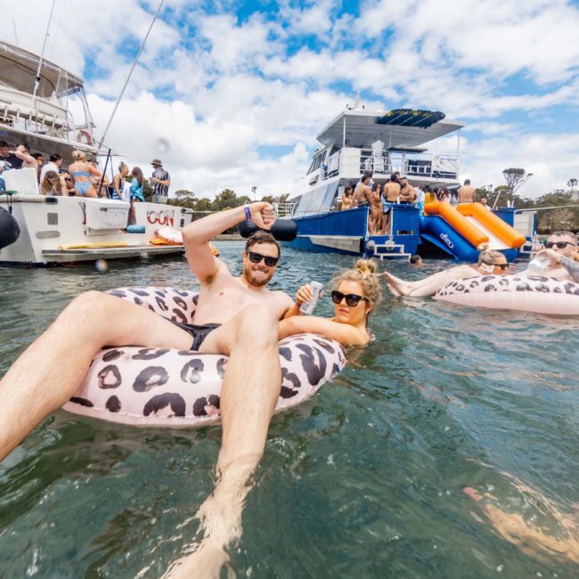 People relaxing on leopard-print floaties in the water near anchored boats, under a partly cloudy sky. Some individuals are on the boats while others swim or float, enjoying a catamaran party Sydney-style.