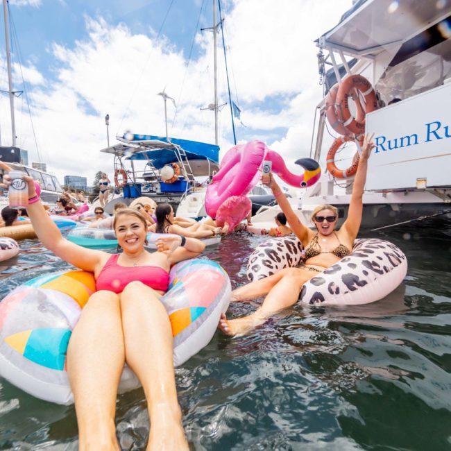 Two women in swimsuits relax on colorful pool floats, raising drinks, surrounded by others in the water near boats on a sunny day during a private yacht charter Sydney Harbour.