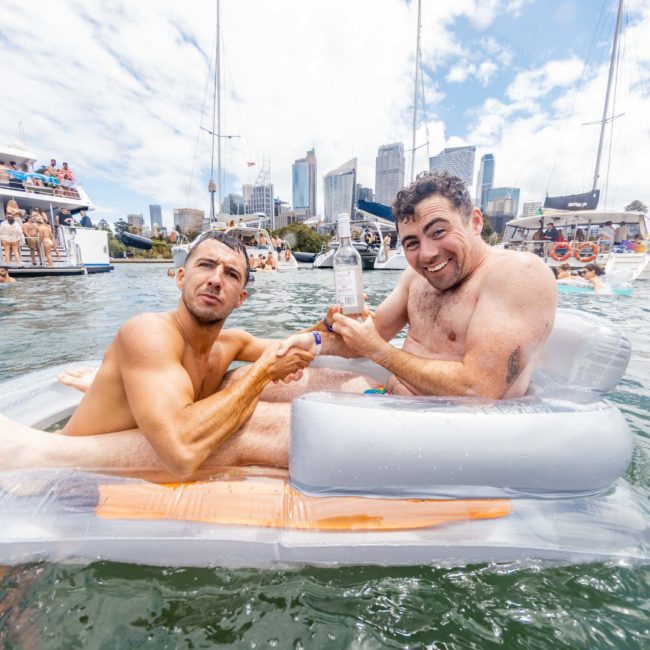 Two men on inflatable loungers shake hands in the water, with boats and a city skyline in the background, adding to the vibe of an exclusive Sydney boat party hire. One man holds a drink bottle.