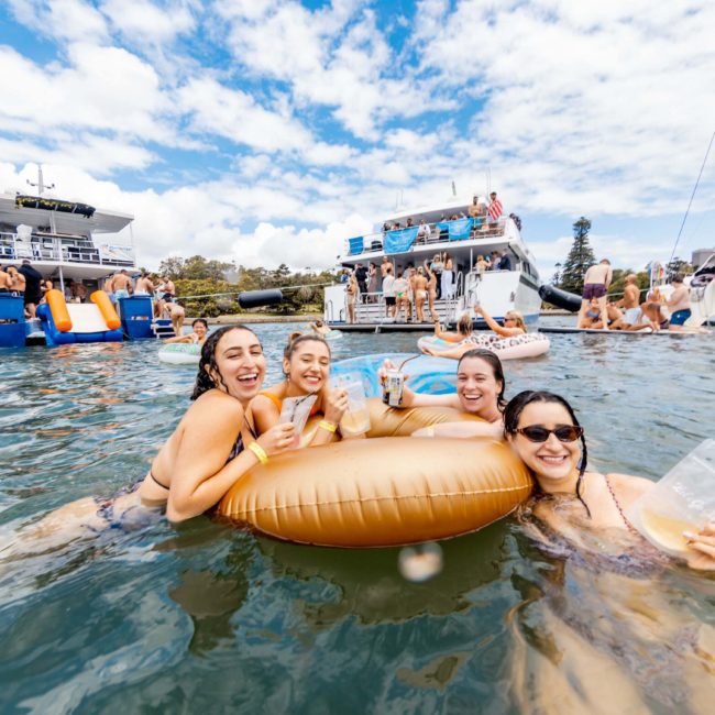 Four women relax on a pool float, smiling and holding drinks amidst a lively Catamaran party in Sydney. Boats and other swimmers can be seen in the background under a partly cloudy sky.