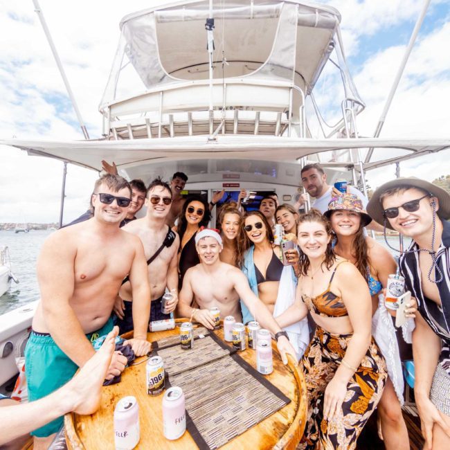 A group of people smiling and posing around a table on a boat, many holding drinks and wearing swimwear. The background features sparkling water and a partly cloudy sky, capturing the essence of a Sydney boat party hire with friends enjoying the moment.