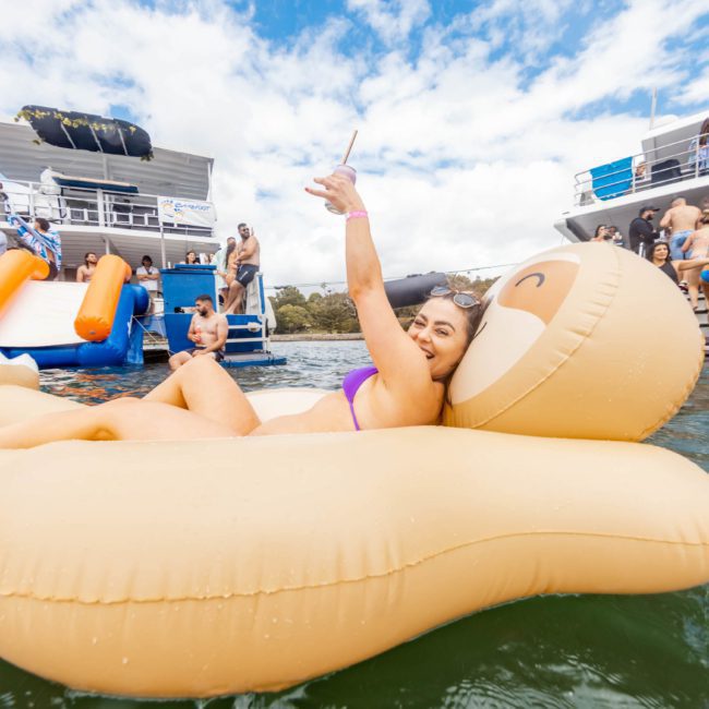 A woman relaxes on an inflatable pool float shaped like a bear, while taking a selfie on a sunny day. In the background, people are on boats and other inflatables, enjoying the water—perfect for a Sydney boat party hire or any corporate boat events Sydney has to offer.