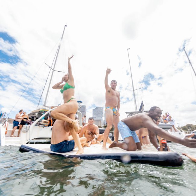 A group of people in swimwear are on a floating platform next to a boat, some jumping into the water and others cheering under a partly cloudy sky during a luxury yacht hire in Sydney Harbour.