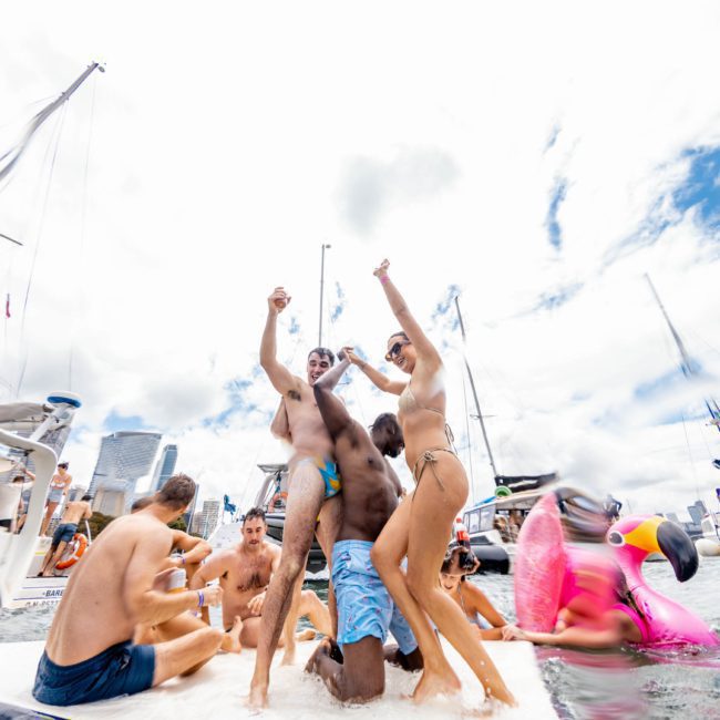 A group of people in swimwear enjoying a party on a boat in a marina, with some dancing and others sitting or floating on inflatables. Multiple boats and a cityscape are visible in the background, embodying the perfect vibe for corporate boat events Sydney.