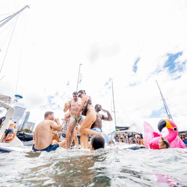 People are gathered on boats and a floating platform near the water with a large pink inflatable bird, under a partly cloudy sky. It's an ideal day for a catamaran party in Sydney.