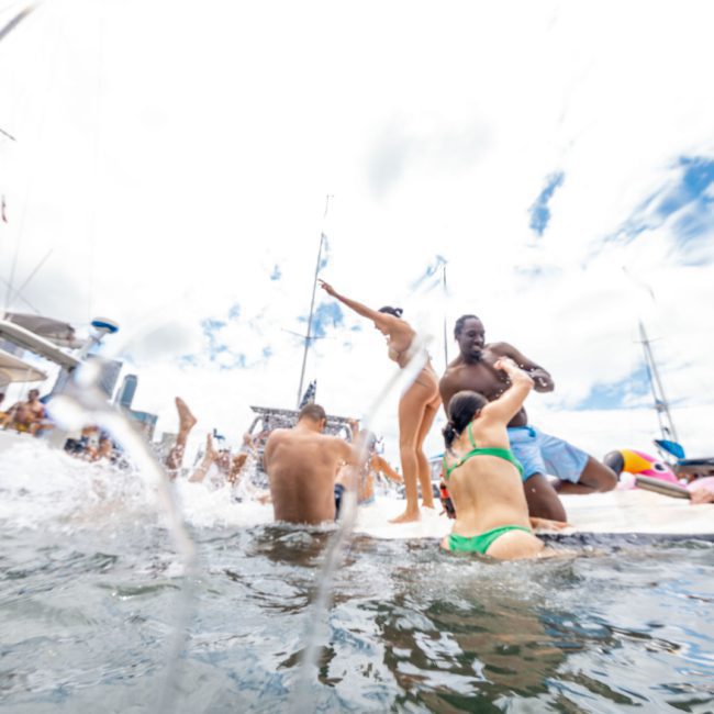 People enjoying a lively time on a boat in the water, dancing and interacting, with splashes and a bright sky in the background, showcasing the fun of luxury yacht hire Sydney.
