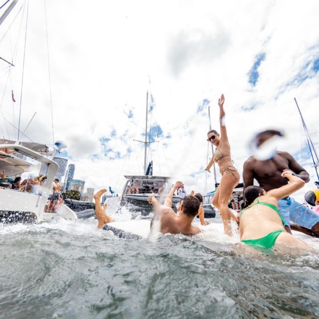 People enjoying a Catamaran party Sydney on a floating platform between boats, with some swimming and others dancing. The scene is festive with a bright and cloudy sky overhead.