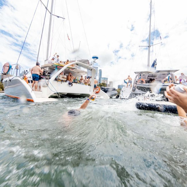 People are swimming near sailboats on a sunny day, with some individuals on the boats enjoying a DJ boat hire in Sydney. The water is splashing, and the cityscape is visible in the background.