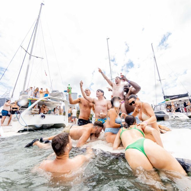 A group of people in swimwear celebrate on a floating platform next to luxury yacht hires in Sydney on a cloudy day. Some are standing and others are seated, with buildings visible in the background, creating the perfect setting for corporate boat events or DJ boat hire parties.