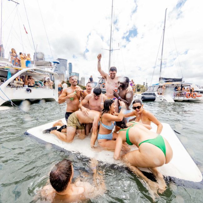 A group of people in swimwear enjoys a party on a floating raft in the water, surrounded by sailboats and yachts under a partly cloudy sky. The catamaran party in Sydney adds an extra layer of excitement to the event.