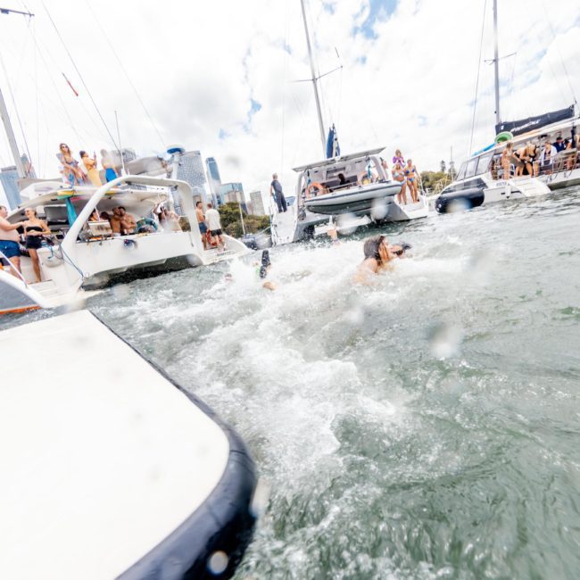 People swimming and engaging in recreational activities around several anchored boats in a bay, enjoying a luxurious yacht hire on Sydney Harbour.