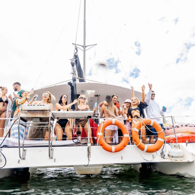A group of people on a boat are enjoying a sunny day. Some are dancing and raising their hands, with life preservers attached to the railings—a perfect scene on this Sydney boat party hire.