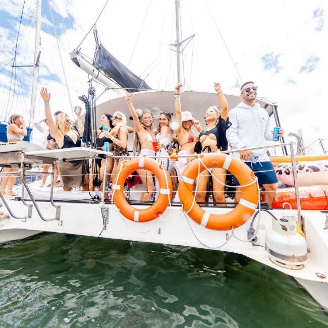 A group of people stand on a boat, some with their arms raised. The boat is on the water with lifebuoys visible under a partly cloudy sky—perfect for corporate boat events in Sydney or an unforgettable catamaran party.