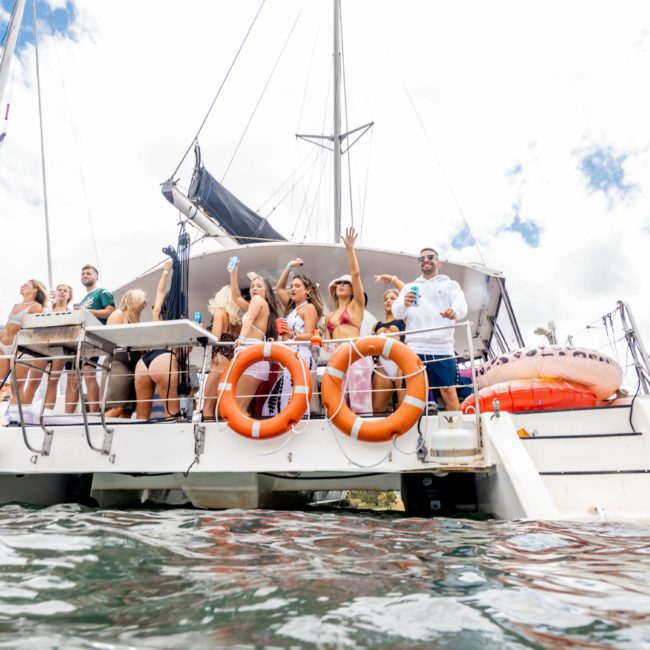 A group of people stand and interact on the deck of a luxury yacht hire Sydney with lifebuoys attached. The sky is partly cloudy, and the yacht is on the water near a shoreline.