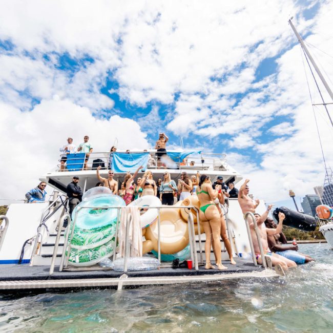 A group of people enjoying a party on a luxury yacht hire Sydney in bright daylight, with large inflatables and a city skyline in the background. Some are on the yacht and others are in the water nearby.