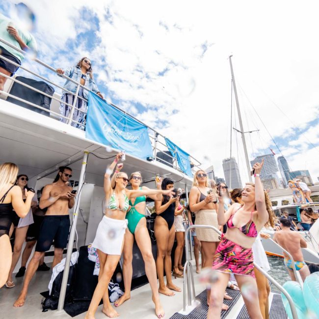 A group of people in swimwear are gathered on a boat during a sunny day. Some are dancing, while others are standing and watching. The background includes a city skyline and a partly cloudy sky, perfect for a Sydney boat party hire.