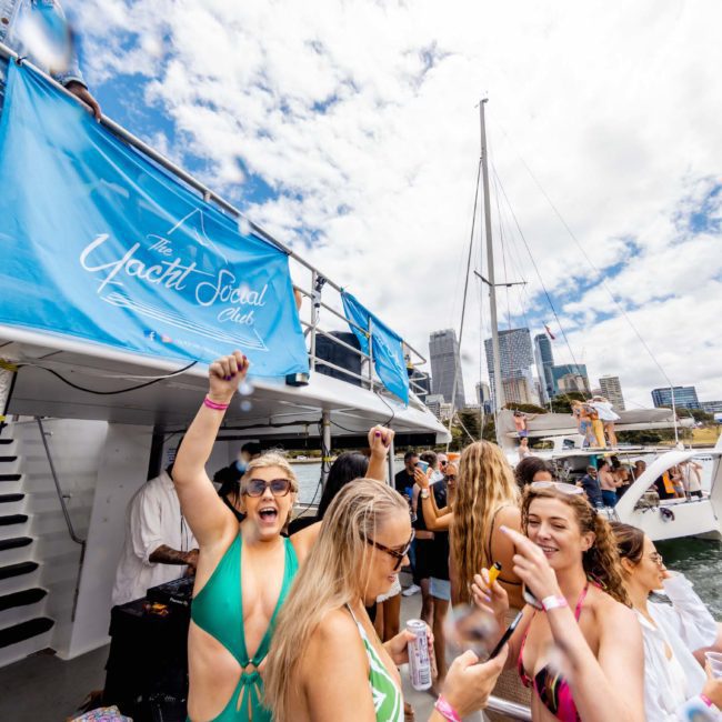 A group of people are partying on a boat with a "Yacht Social Club" banner. Boats and a city skyline are visible in the background under a partly cloudy sky, perfect for your next Sydney boat party hire or private yacht charter on Sydney Harbour.